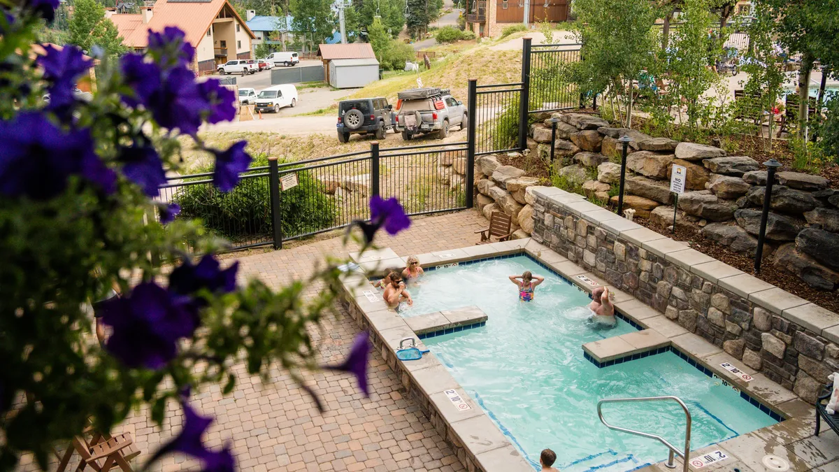 A vibrant swimming pool surrounded by stone walls and flowering plants, with children and adults playing and relaxing in the water. The area is sunlit and lively, evoking a cheerful atmosphere suitable for family fun or tranquil swims at the Purgatory Lodge.