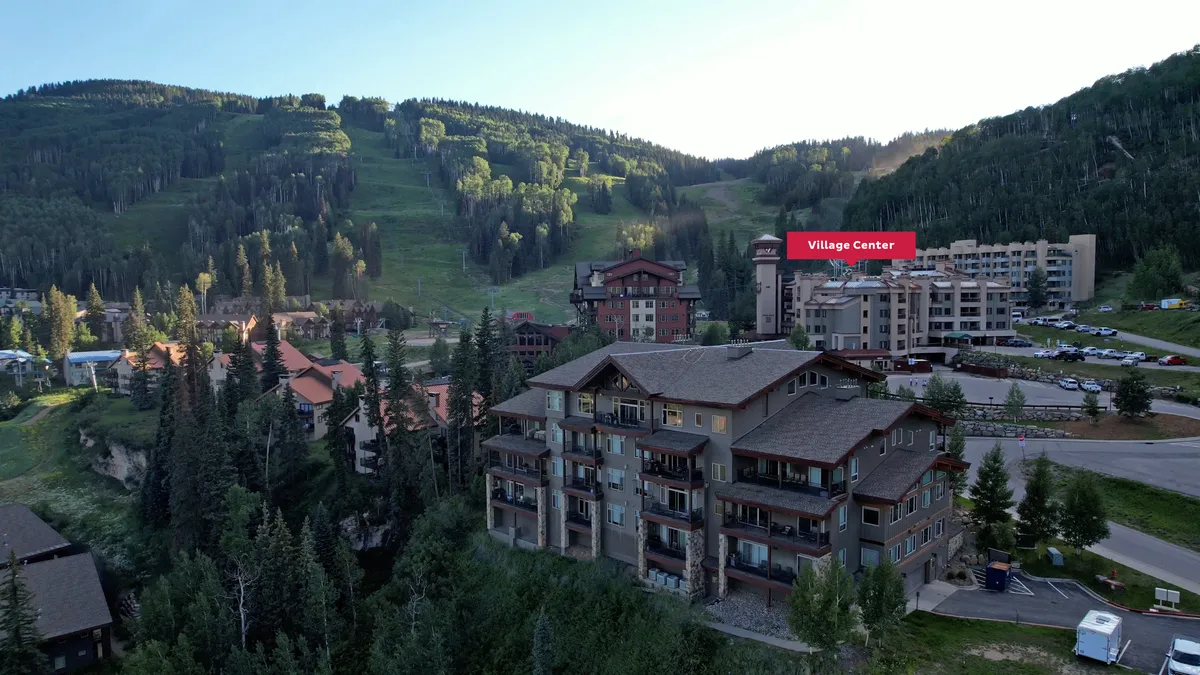 A multi-story mountain resort building sits nestled among tall trees, framed by a stunning mountain backdrop. The ski slopes are clearly visible beside the building, showcasing both the adventure and natural beauty of the location. The scene exudes a tranquil yet adventurous mood, inviting guests to enjoy both relaxation and outdoor activities.
