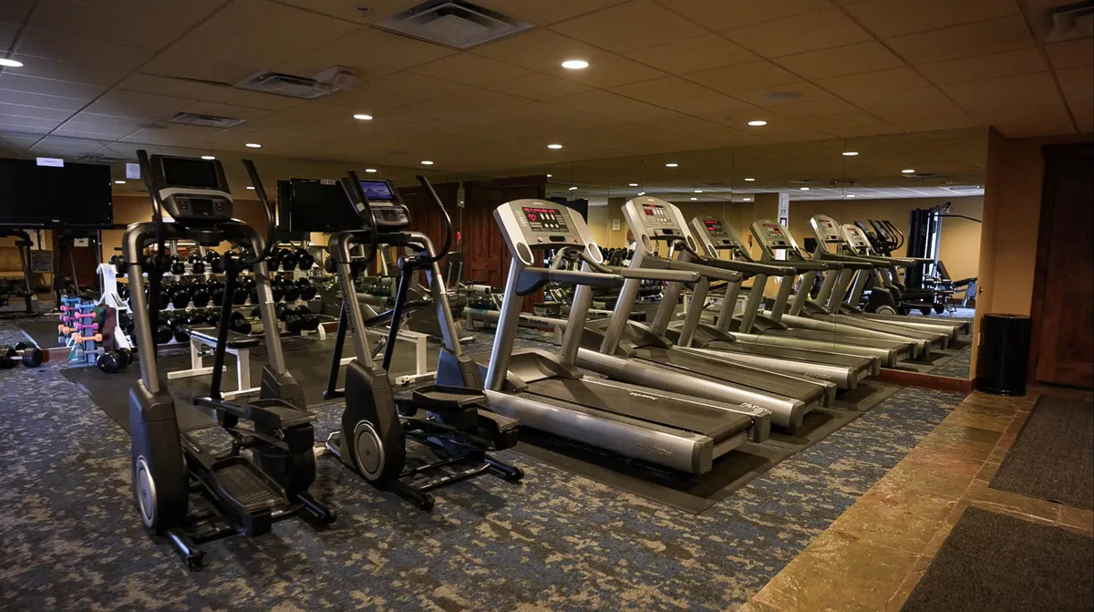 An interior fitness center equipped with treadmills, ellipticals, dumbbells, and a mirrored wall. The space is bright, modern, and inviting, set up for both individual workouts and group classes, reflecting a commitment to health and wellness.