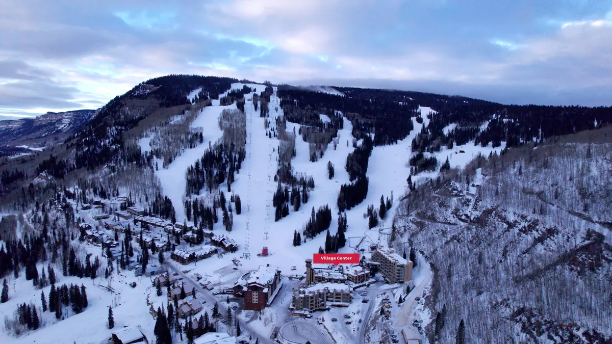 A vibrant exterior view of the Village Center, with snow-dusted rooftops, ski slopes bustling with activity, and a lively mountain village atmosphere. The surrounding trees and mountains create a stunning backdrop, and the image captures skiers and visitors enjoying the convenience of slope-side access and village amenities.