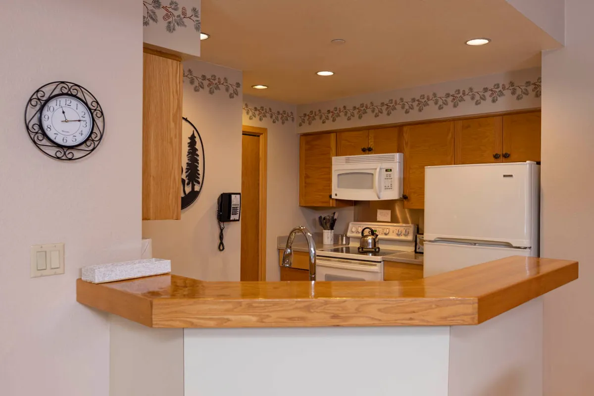 A cozy kitchen scene with a central kitchen island, warm wooden cabinets, white appliances, and decorative wall trim. The space is inviting and well-lit, with a visible clock adding to the sense of homeliness. The kitchen island offers ample workspace for meal preparation, while the overall layout encourages social interaction during cooking and entertaining.