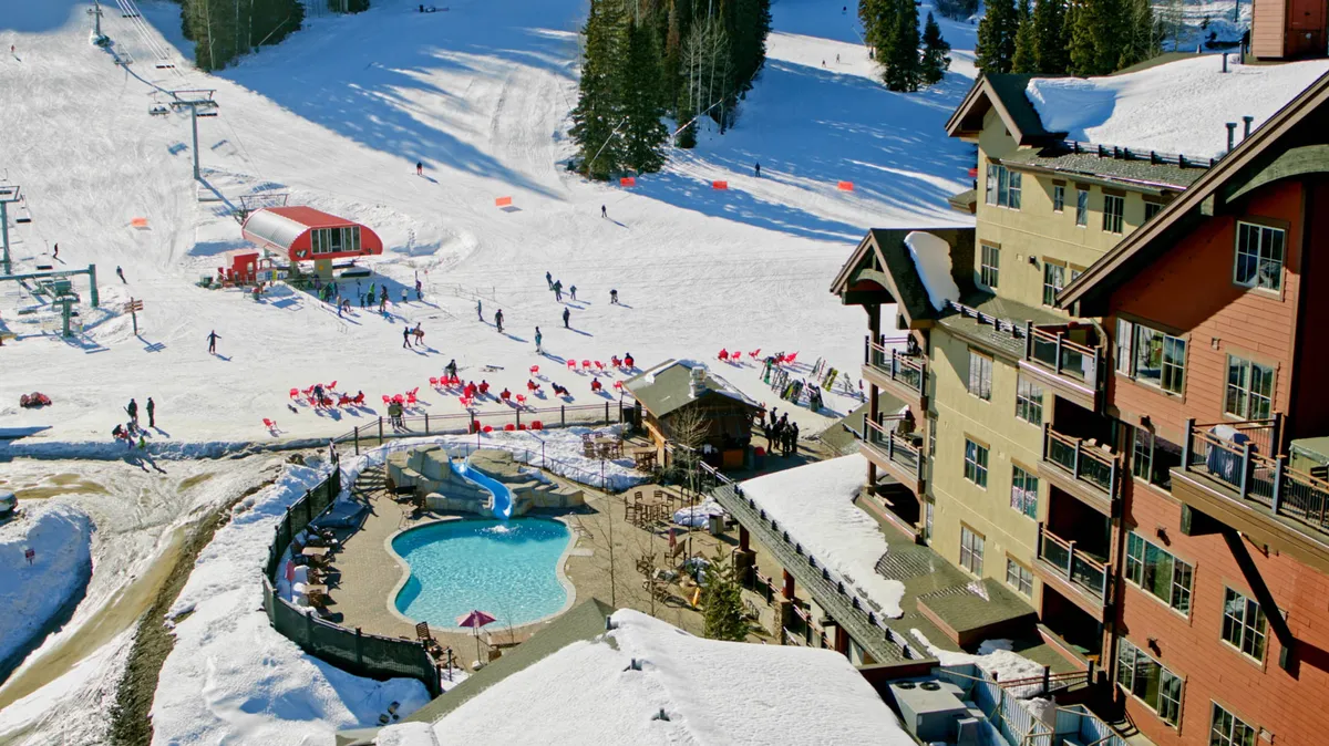 The snowy ski slopes are alive with families and skiers, adjacent to an outdoor pool and a welcoming lodge building. The image captures people skiing, children playing in the snow, and guests relaxing by the pool, all set against a stunning alpine backdrop.