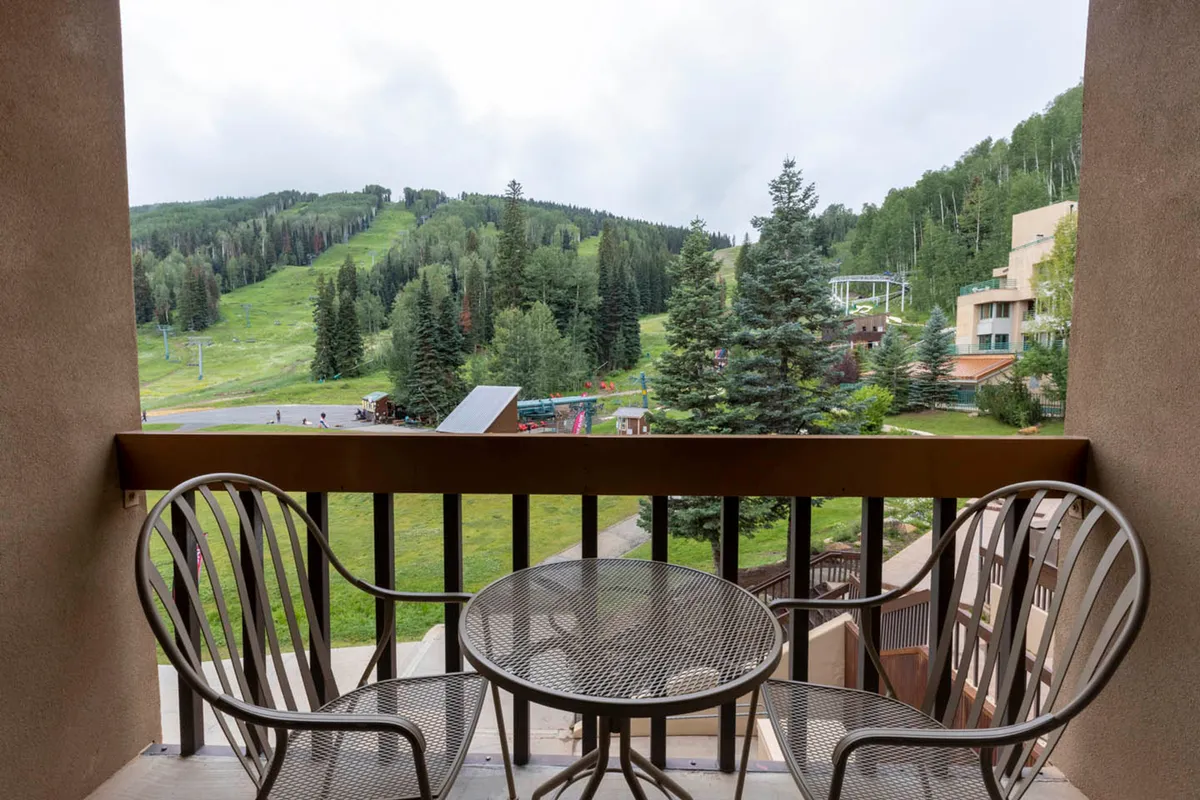 A cozy balcony with comfortable seating overlooks a panoramic mountain view surrounded by trees. The tranquil atmosphere invites guests to relax after a day of skiing, catch a sunrise, or stargaze from the comfort of their condo.
