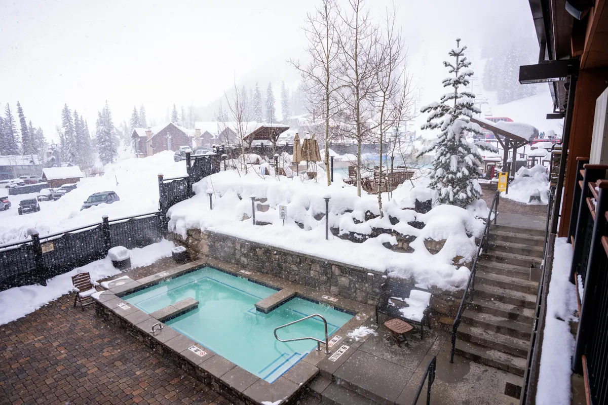 An exterior hot tub surrounded by a snow-covered landscape and tall trees, with steep stairs leading down to the area. Warm steam rises from the bubbling water, creating a stark yet inviting contrast with the wintry scenery. The ambience is peaceful and perfectly suited for relaxation after a day outdoors.