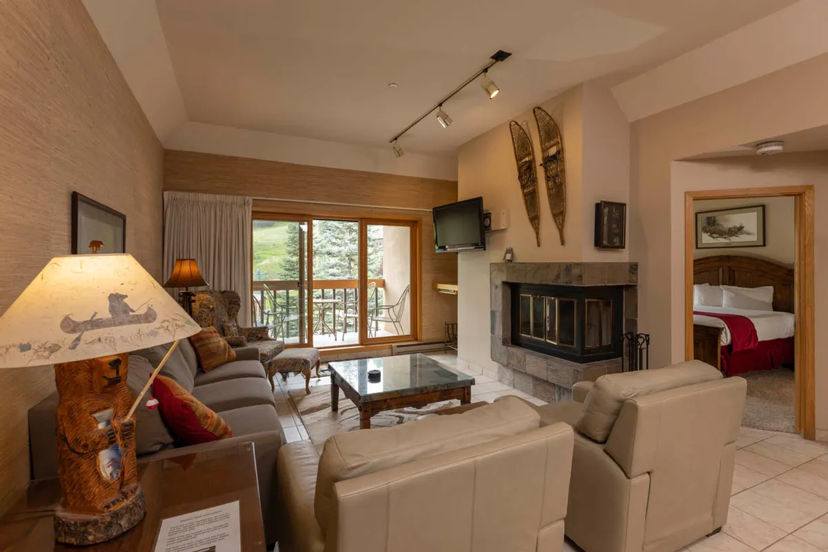 The image showcases a warm, inviting living area featuring a modern fireplace, a plush sofa set perfect for gathering, large windows framing breathtaking mountain vistas, and artistic touches like a sculptural lamp and wood accents. The space exudes a cozy, mountain-lodge ambiance, with the golden light of sunset streaming in and the promise of comfort and relaxation.