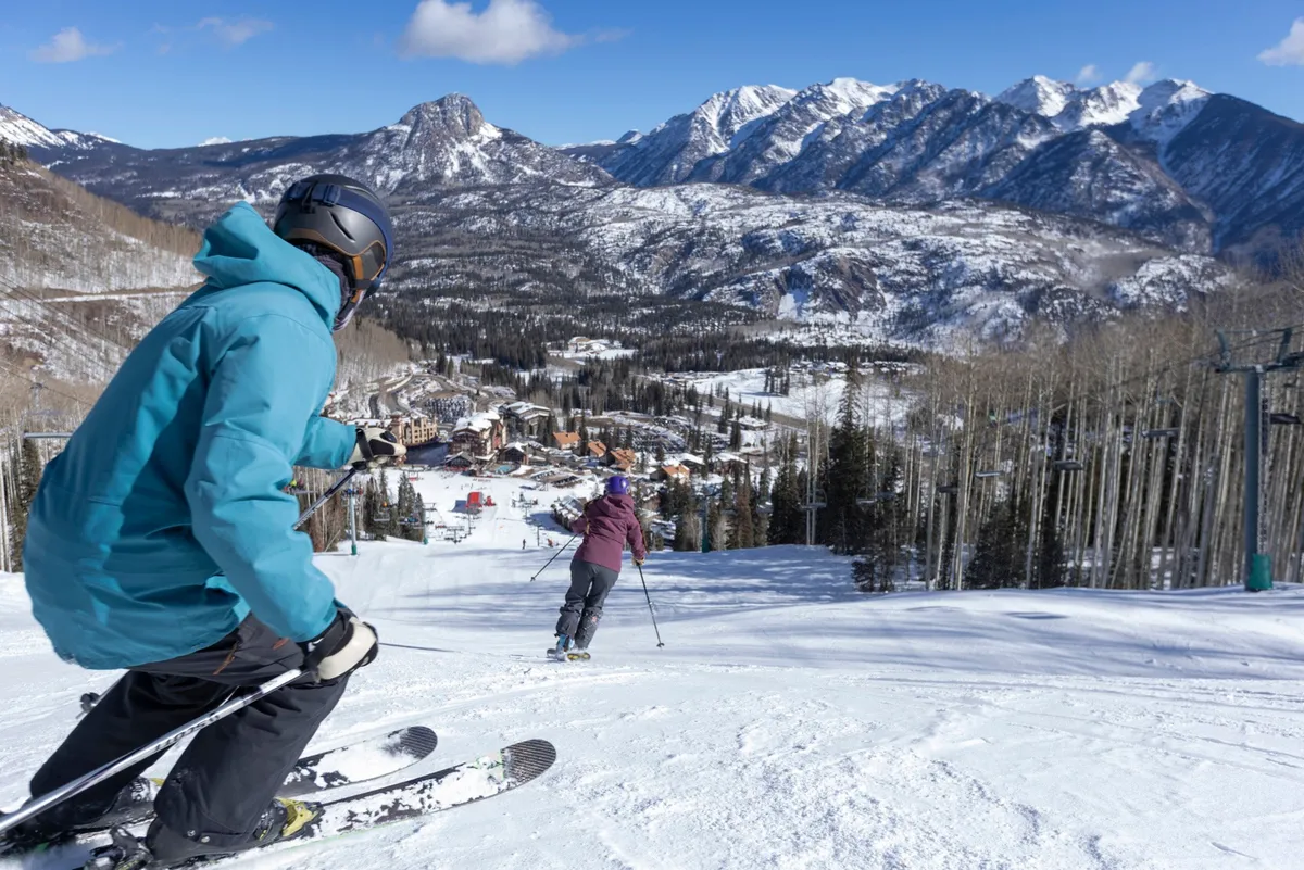 A dynamic winter scene showing skiers descending snowy slopes with majestic mountains rising in the background and resort buildings nestled at the base. The energetic atmosphere captures the thrill and camaraderie of group ski adventures.
