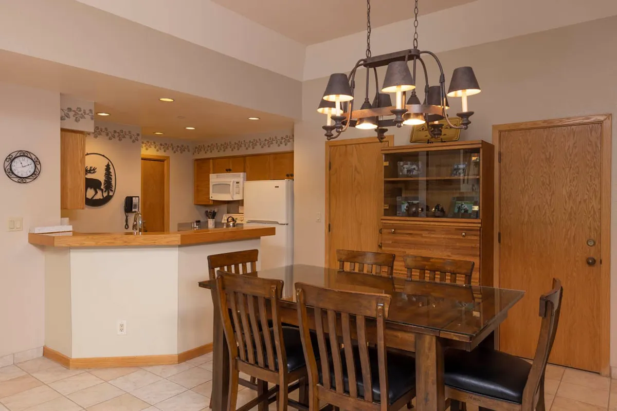 A communal dining area with a large table and comfortable chairs, illuminated by a striking chandelier. The kitchen counter in the background signals an open-concept space perfect for entertaining or family meals, reinforcing the idea of shared experiences in the heart of the home.