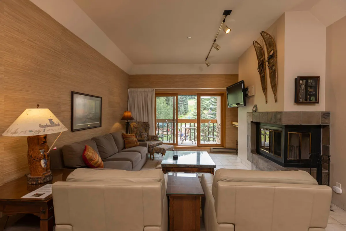 A warm, inviting living room featuring a plush sofa arranged around a modern fireplace. Large windows frame expansive mountain views, and soft lighting from stylish lamps enhances the cozy atmosphere. The scene suggests a place to relax, socialize, or watch TV while staying connected to the scenic outdoors.