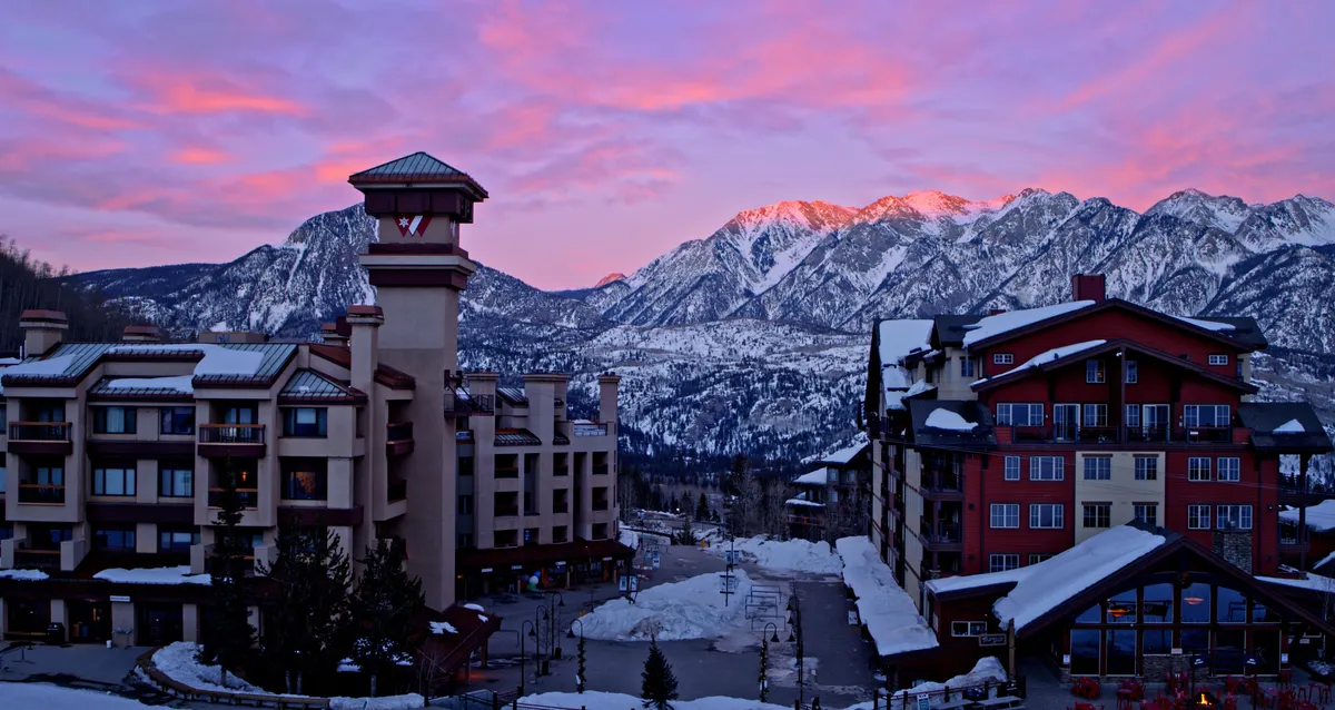 A stunning exterior view of the Needles Mountain Range with snow-capped peaks, sunlight casting a warm glow over the landscape, and cozy buildings nestled at the base. The scene conveys a sense of tranquility and awe, perfect for highlighting the breathtaking mountain setting.