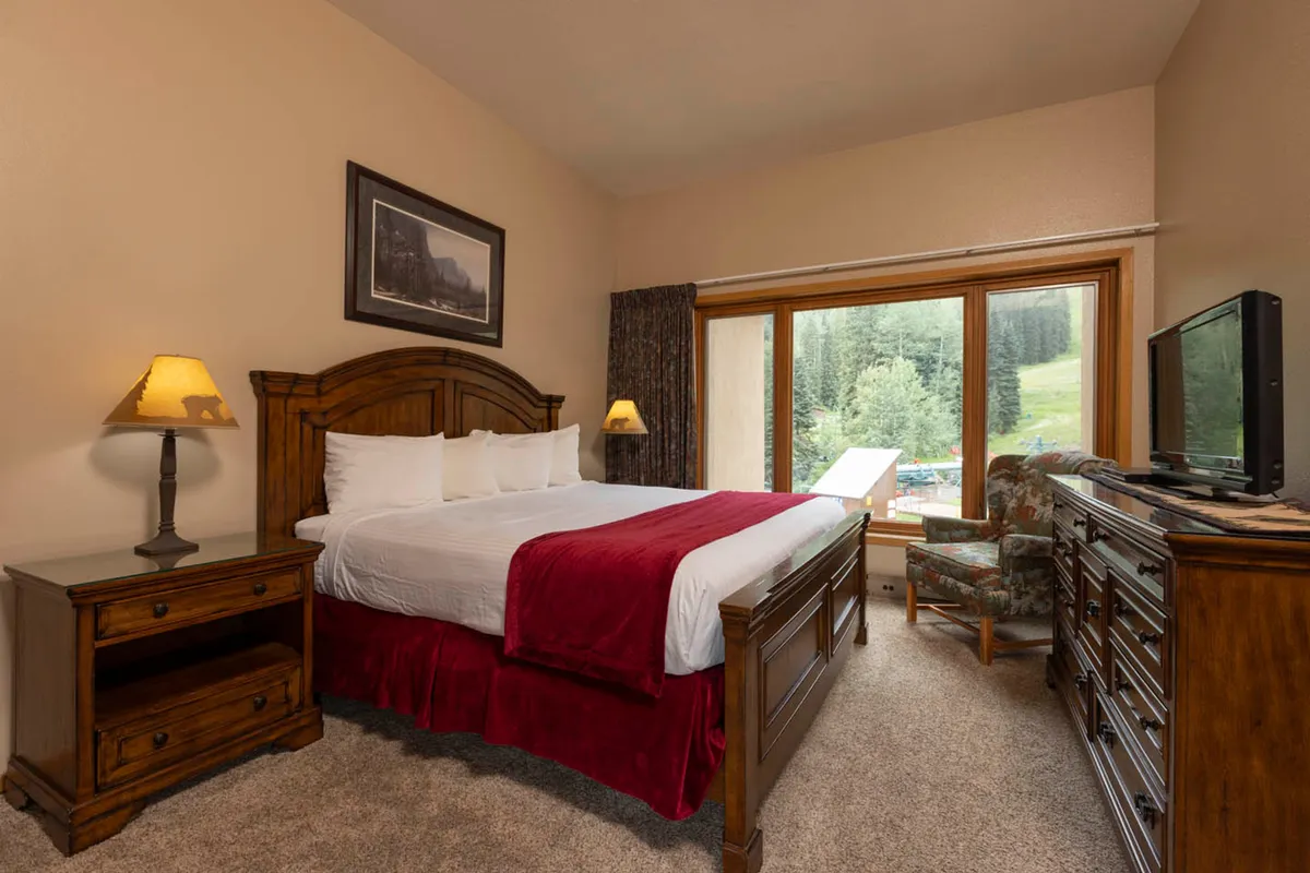 A bright, serene bedroom with a king-sized bed dressed in white linens and a red throw blanket. A large window frames beautiful mountain scenery, while wooden nightstands and a reading chair create a welcoming, tranquil space.