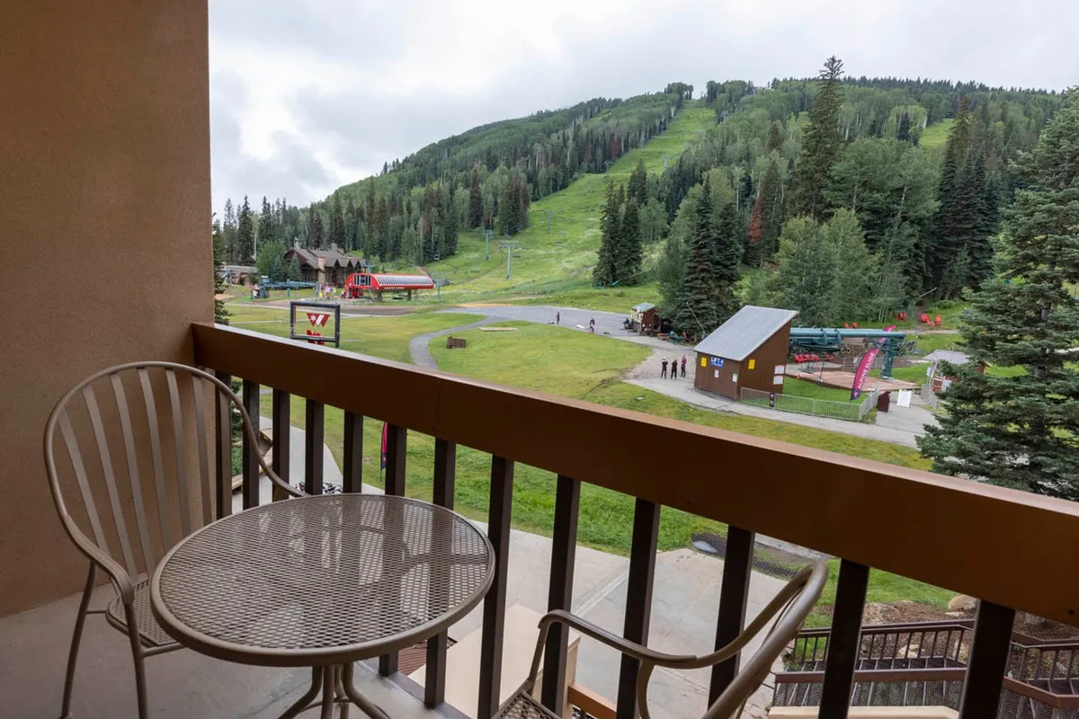 A scenic balcony scene featuring comfortable chairs overlooking a breathtaking mountain landscape, with the ski lift and nearby pathways in view. The vista includes snow-capped peaks, suggesting effortless access to skiing and relaxation amid natural beauty.