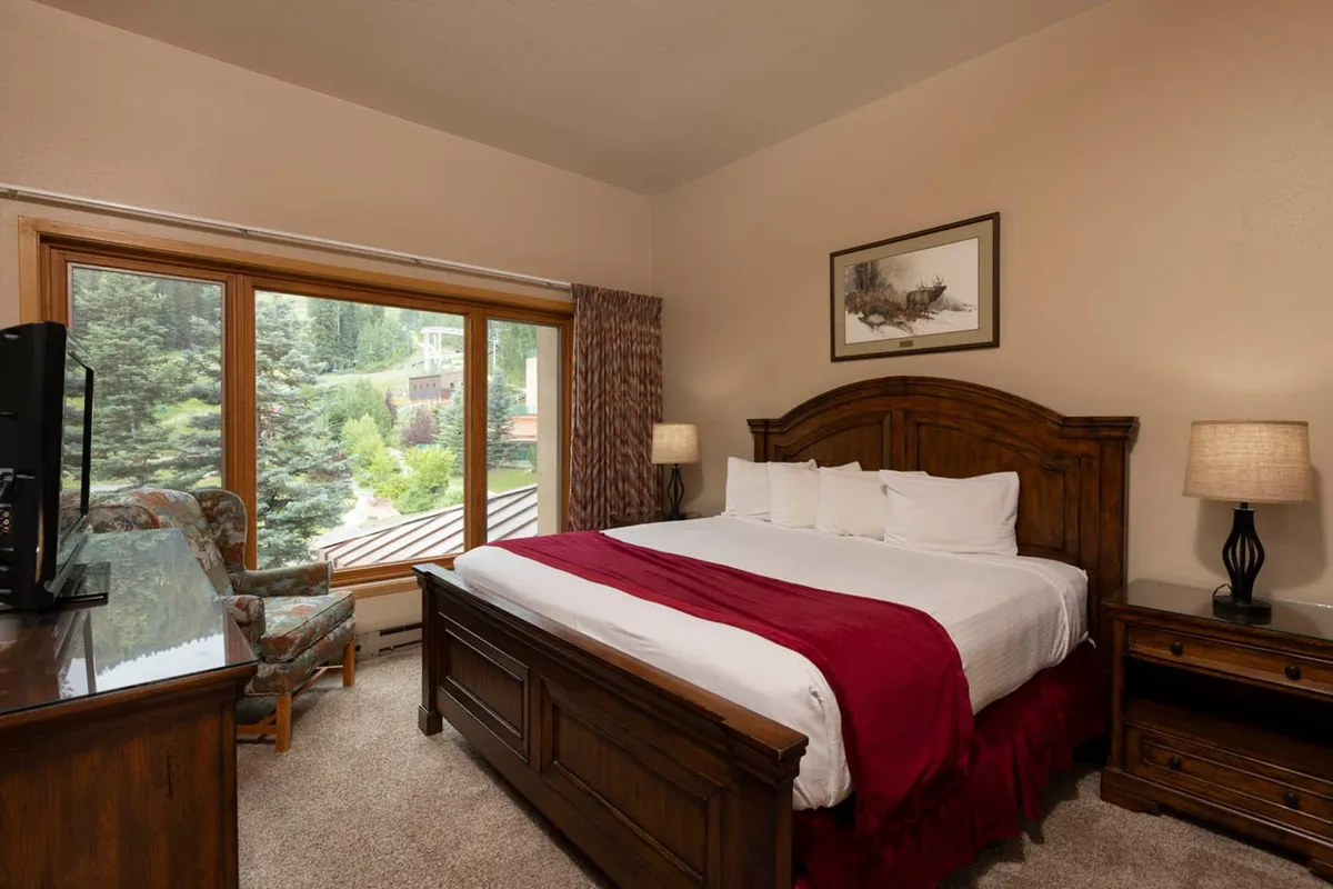 A spacious bedroom featuring a large bed adorned with white linens and a red throw blanket. The room includes large windows that let in ample natural light and offer scenic mountain views. Warm wooden furniture adds to the inviting and restful atmosphere.